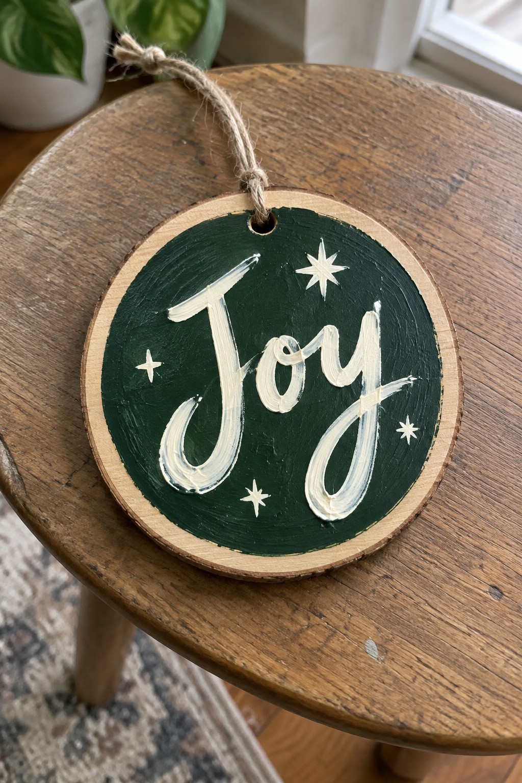 Circular green wooden ornament painted with white cursive Joy, stars, and twine hanger.