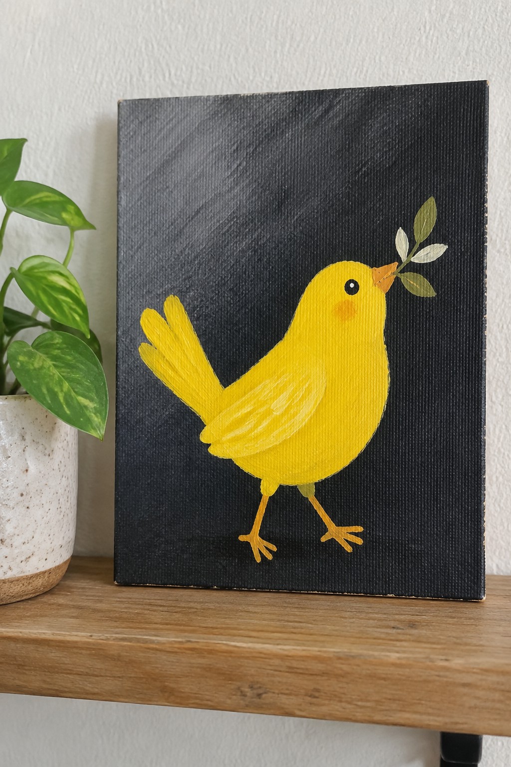 Yellow bird painting on black canvas beside potted plant on wooden shelf