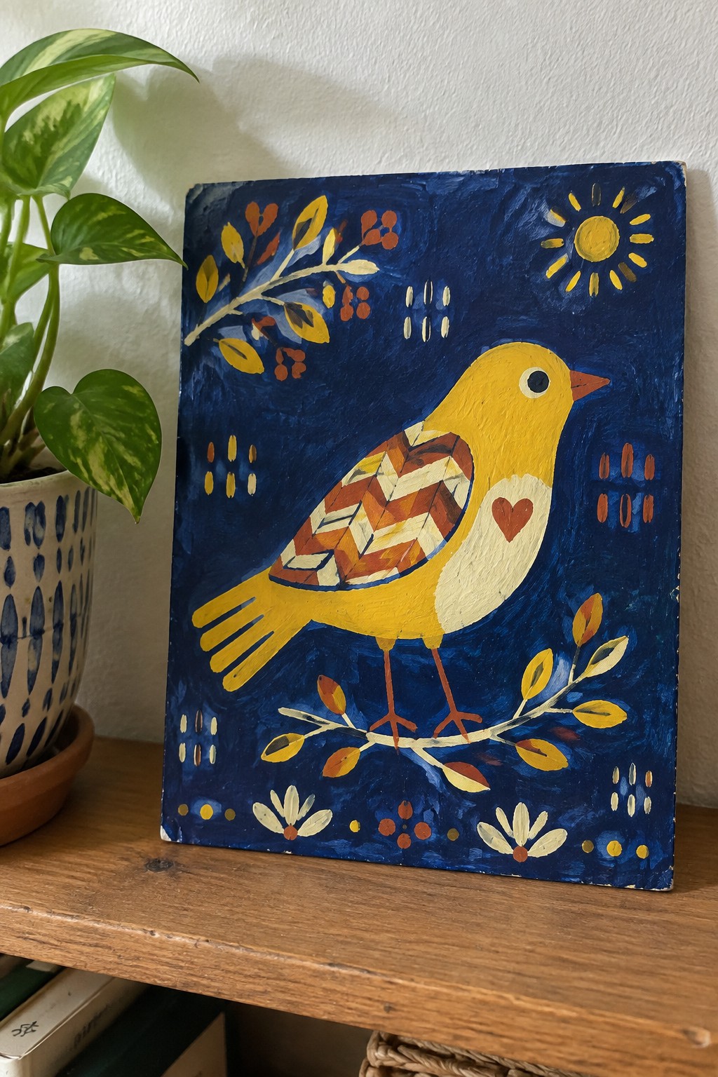 Folk art painting of yellow bird with patterned wings and heart on blue background beside plant.
