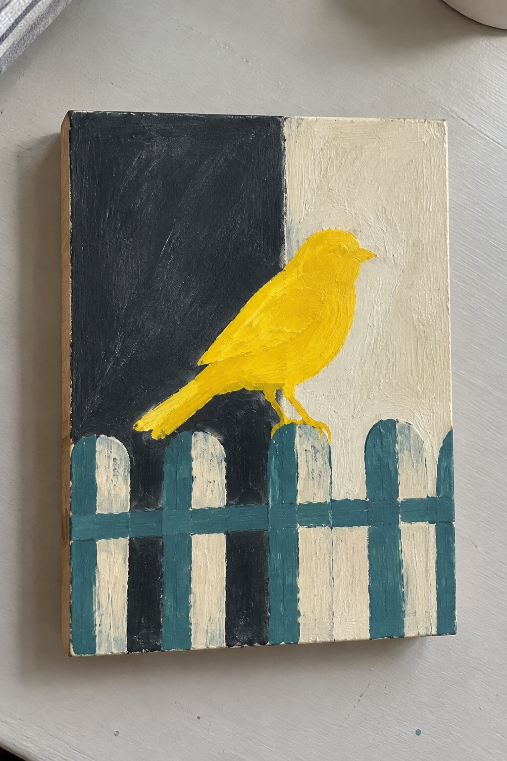 Yellow bird perches on teal picket fence against split black-and-white background.