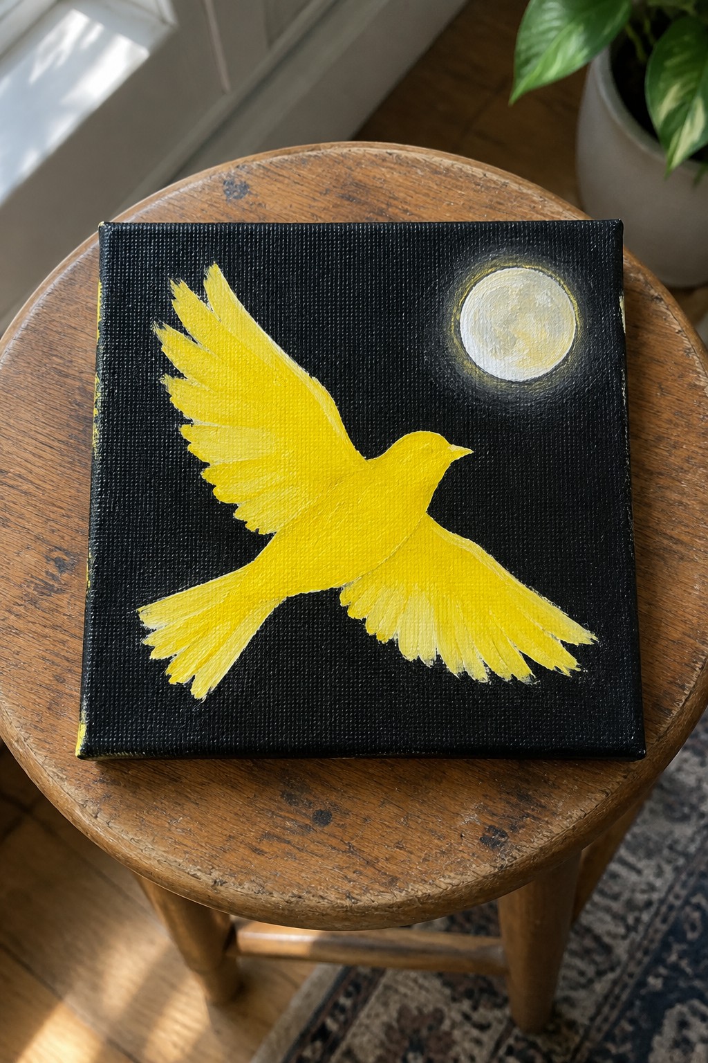 Yellow bird painting on black canvas with moon, atop wooden stool.