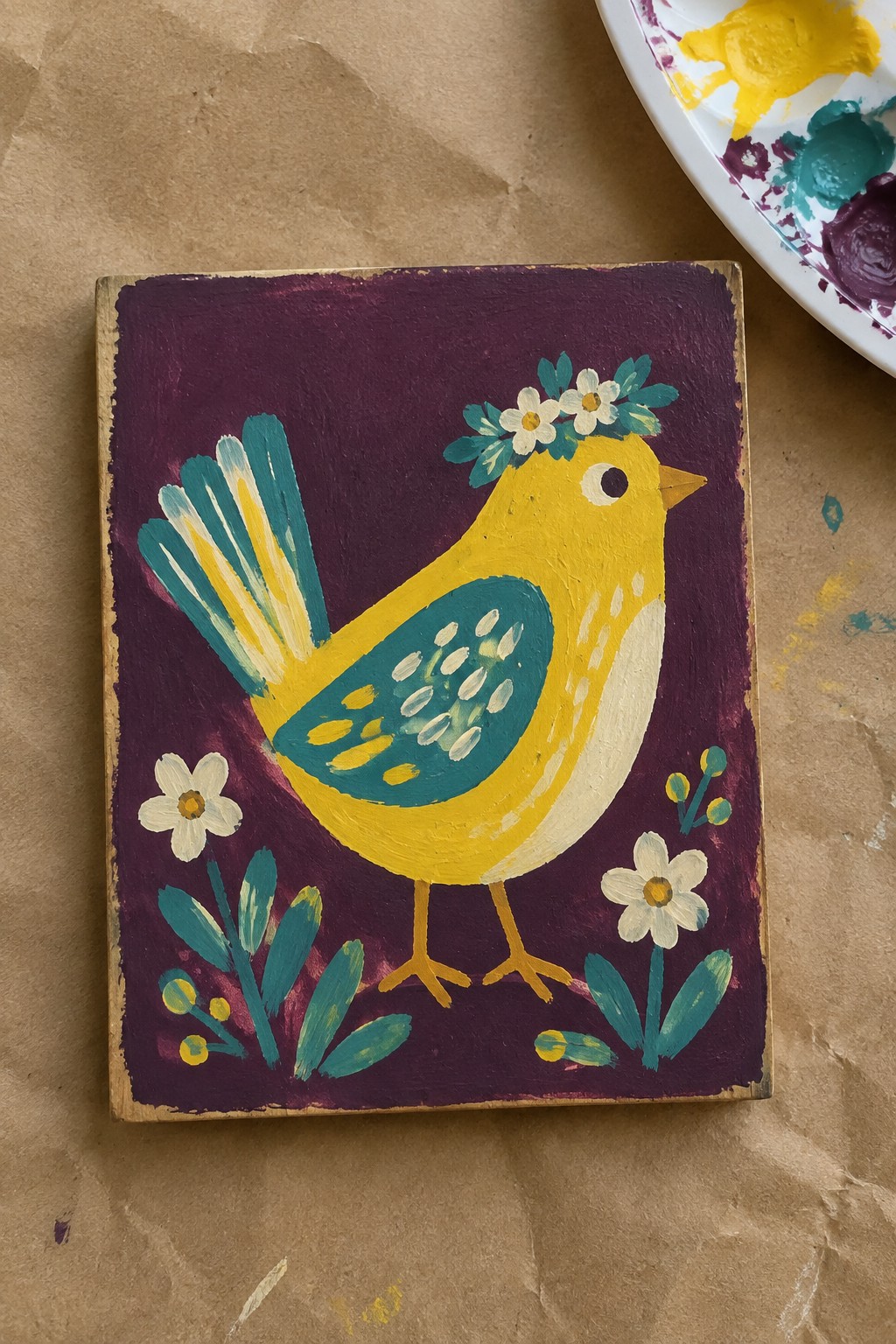 Acrylic painting of a yellow bird with a flower crown on a purple background with flowers and leaves at the bottom.