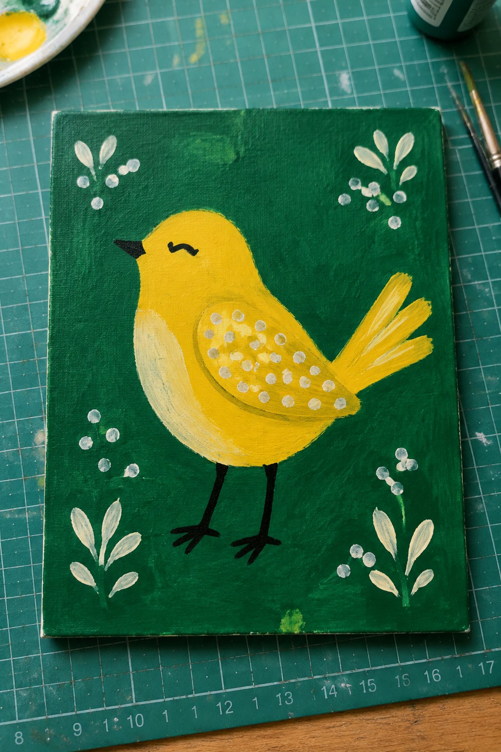 Yellow bird with dotted wing on green canvas amid white foliage and art supplies