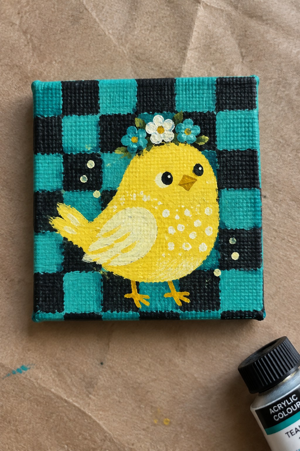 Yellow chick with flower crown on teal black checkered canvas beside paint tube