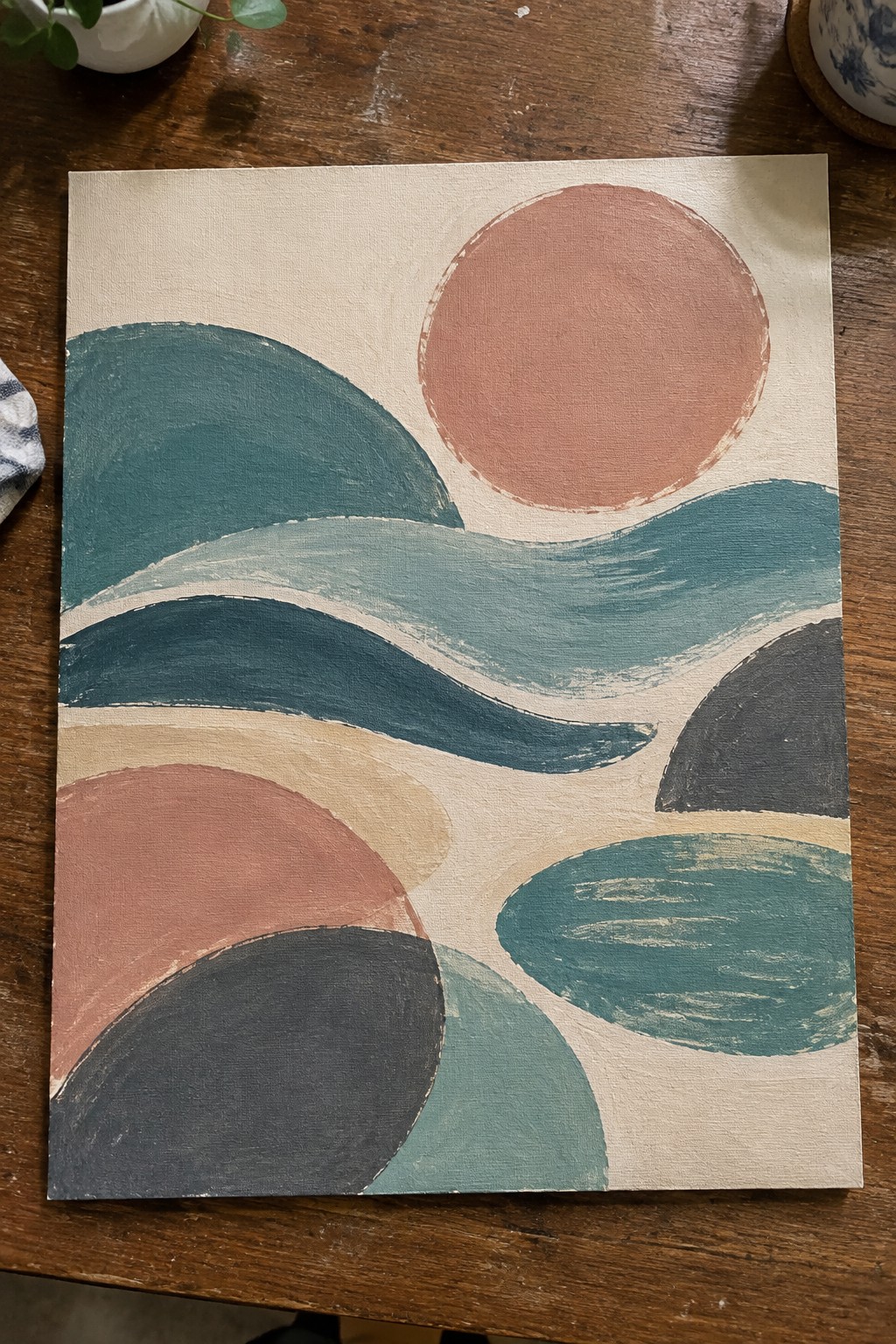 Abstract canvas with teal waves, coral circles, and dark organic shapes on wood table