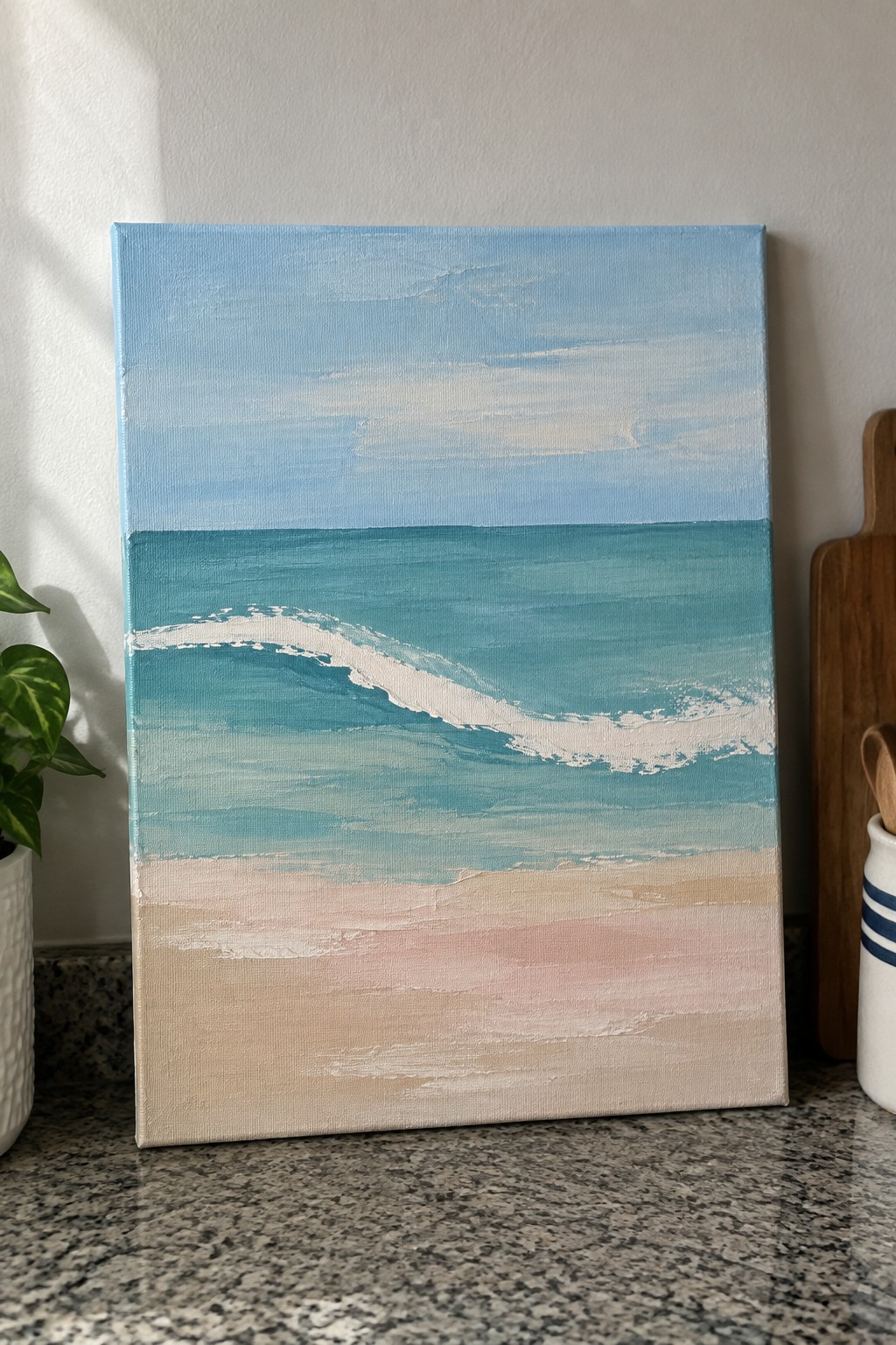 Canvas beach painting with blue wave on granite counter beside potted plant