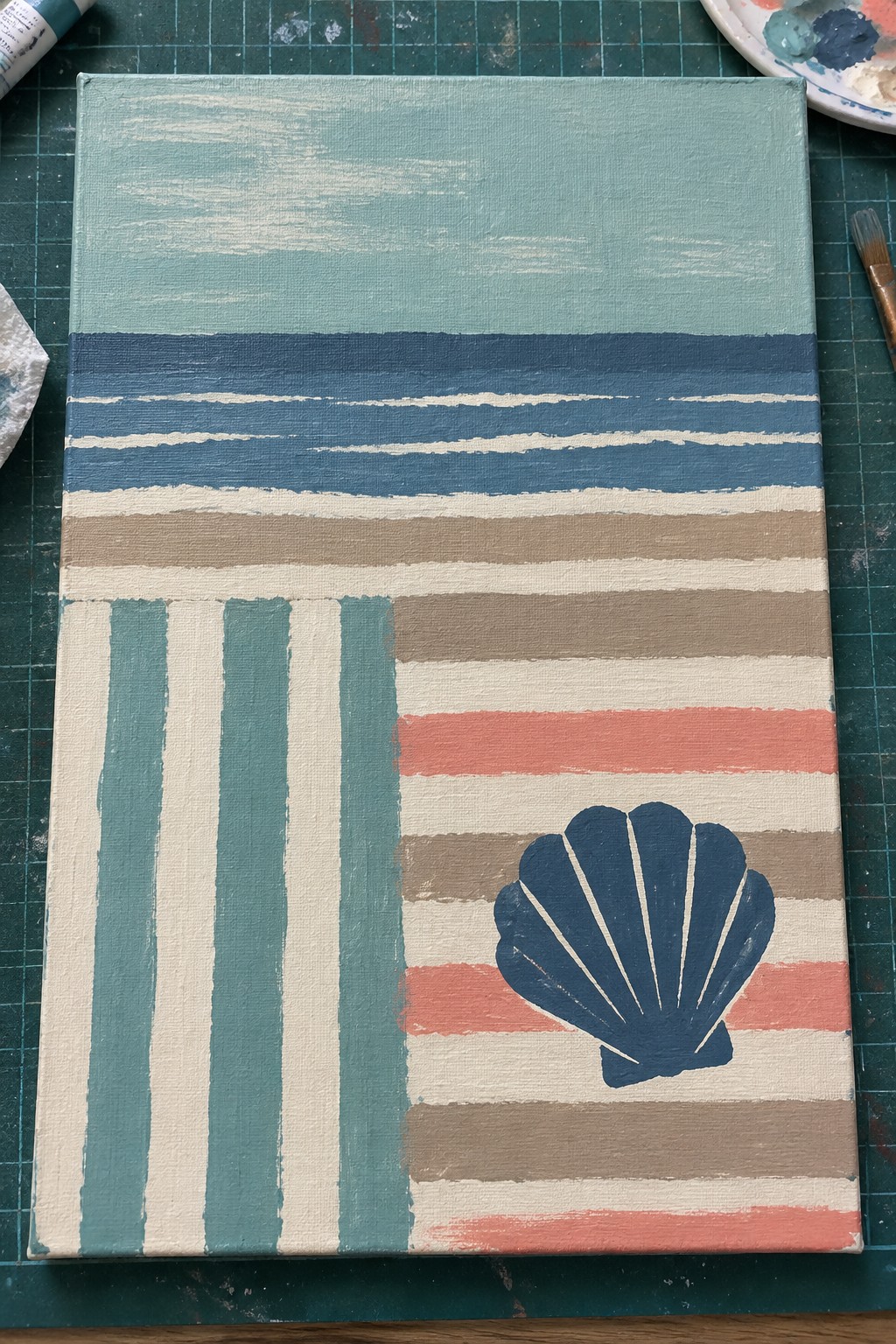 Abstract canvas with colorful stripes and blue seashell on green cutting mat with paints