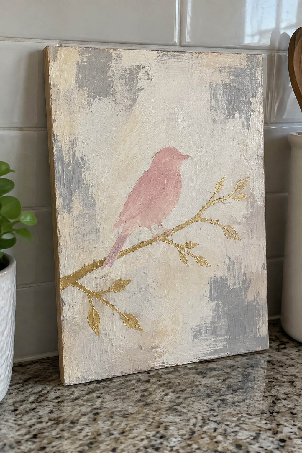 Pink bird on gold branch against textured beige-gray distressed canvas on countertop.