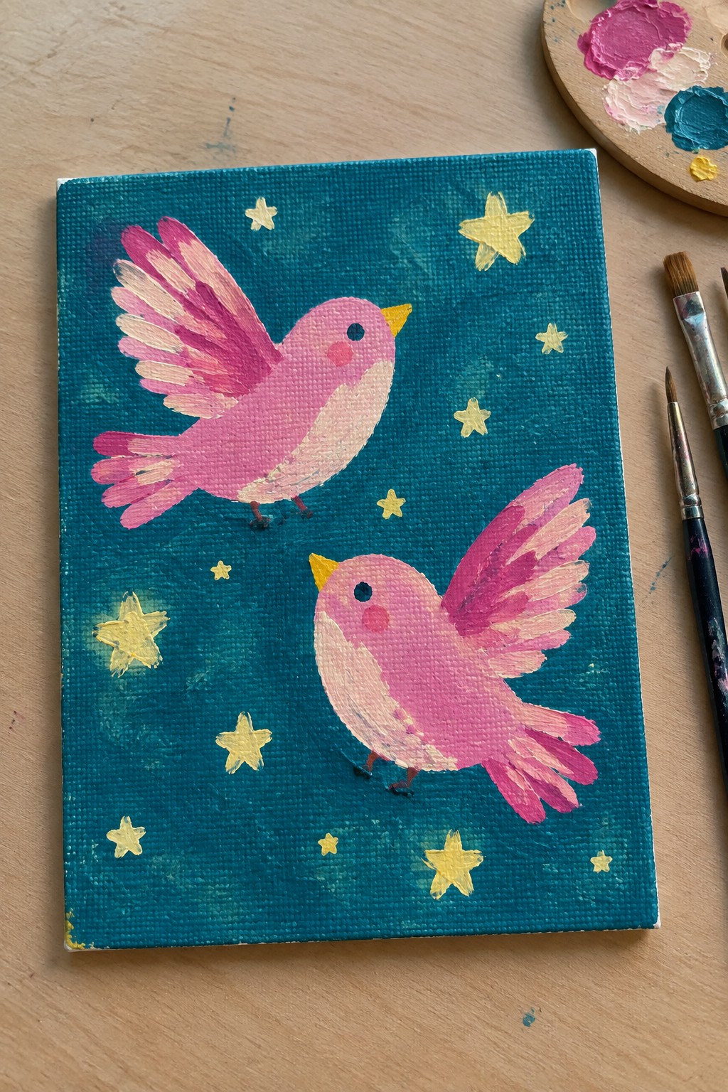 Two pink birds soaring on teal canvas amid yellow stars with paint palette and brushes