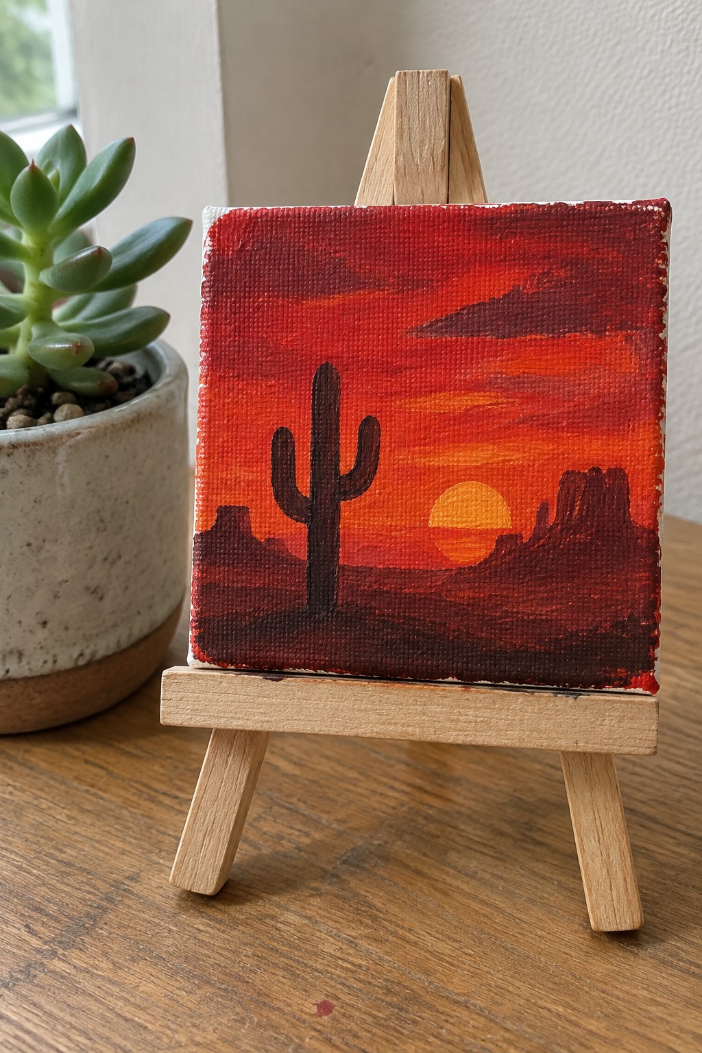 Miniature desert sunset painting with cactus on wooden easel beside succulent plant