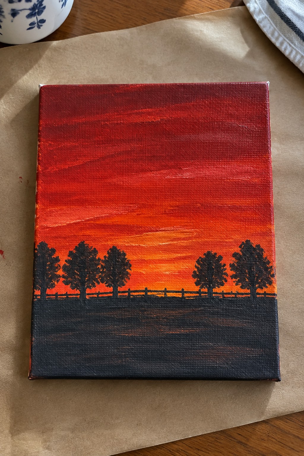 Canvas painting of vibrant red-orange sunset with black tree silhouettes and fence.