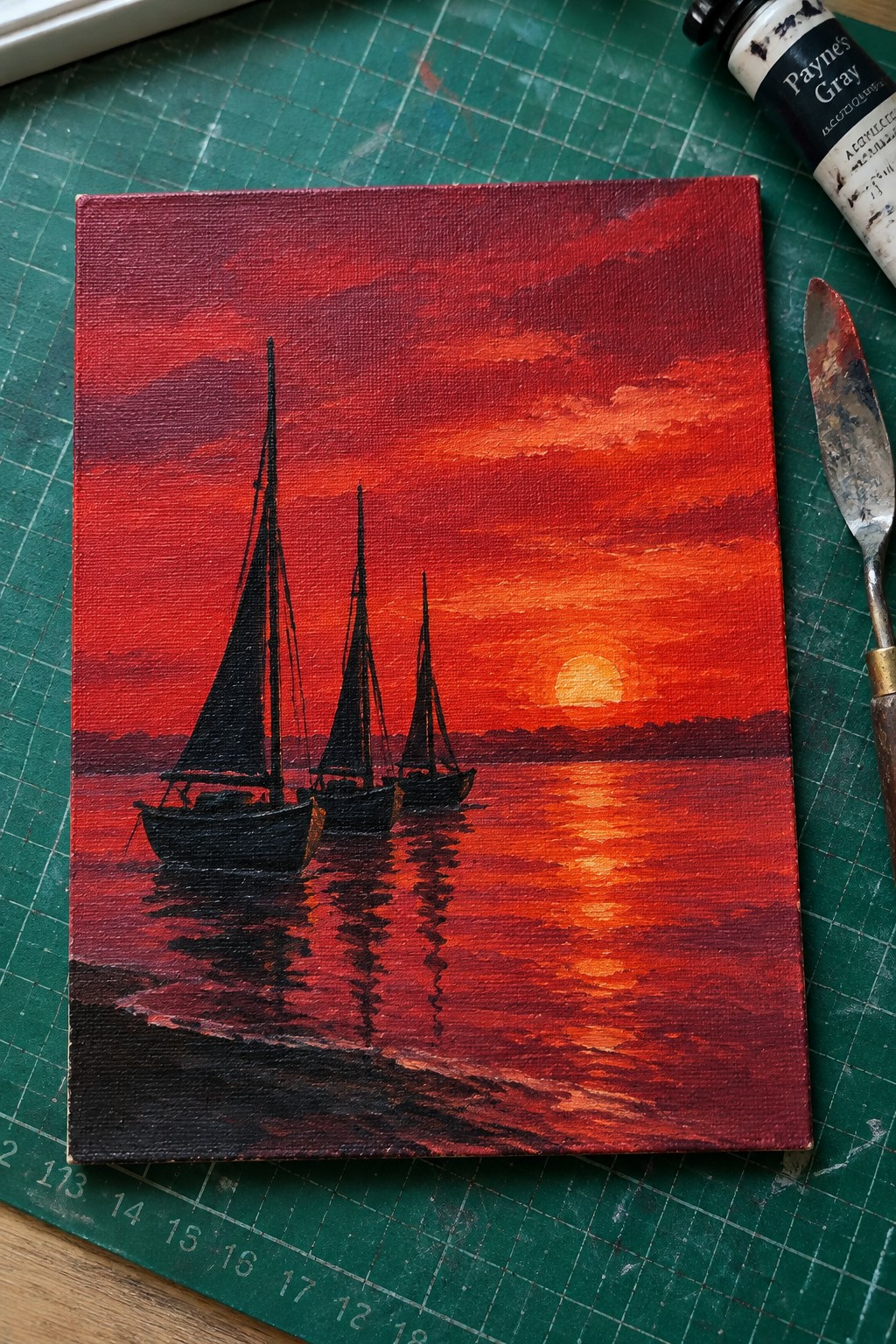 Three black sailboats on reflective water under vivid red sunset sky with paint tube and knife nearby
