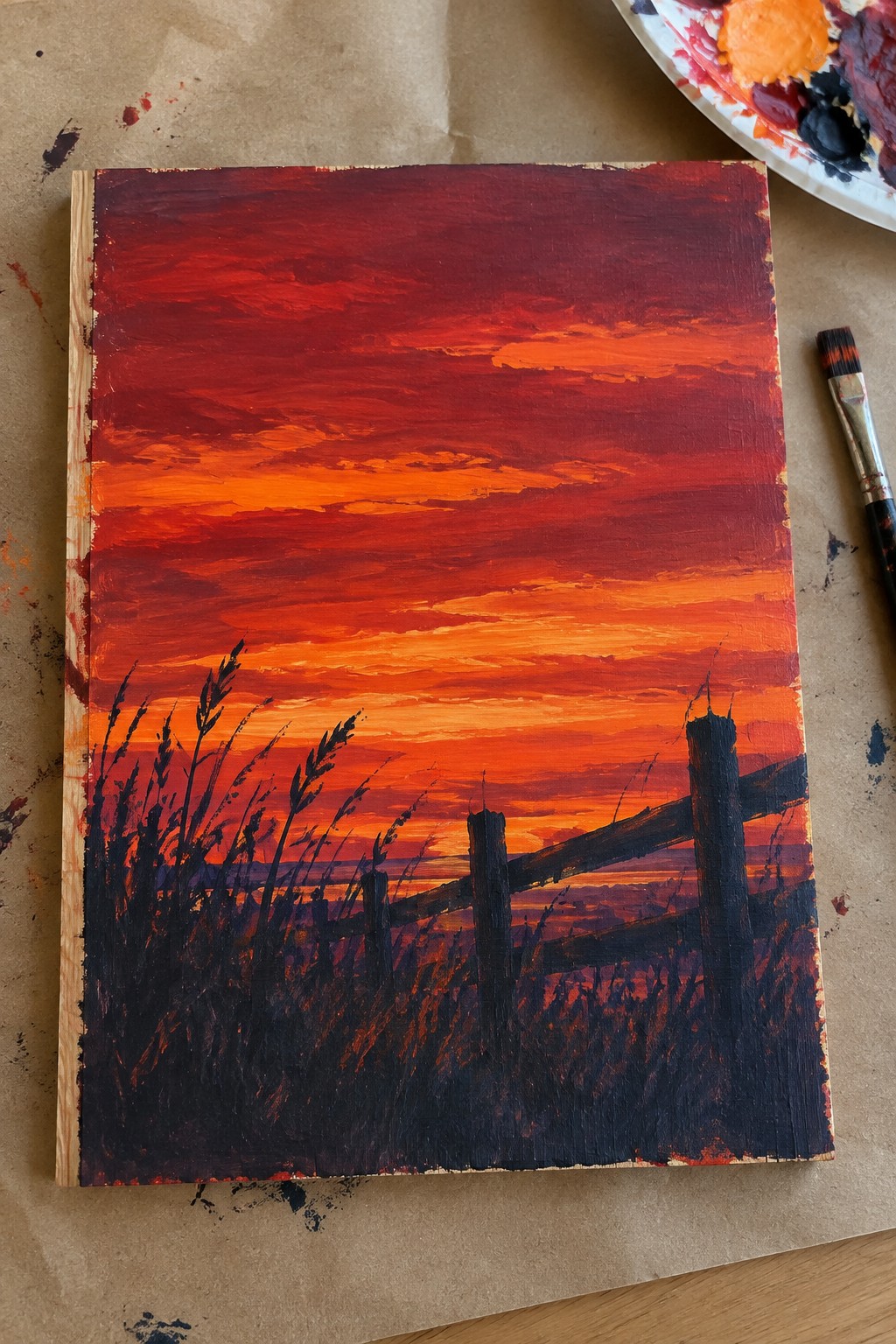An acrylic painting of a red-orange sunset sky with horizontal brushstrokes, a dark wooden fence, and tall grass silhouettes in the foreground.