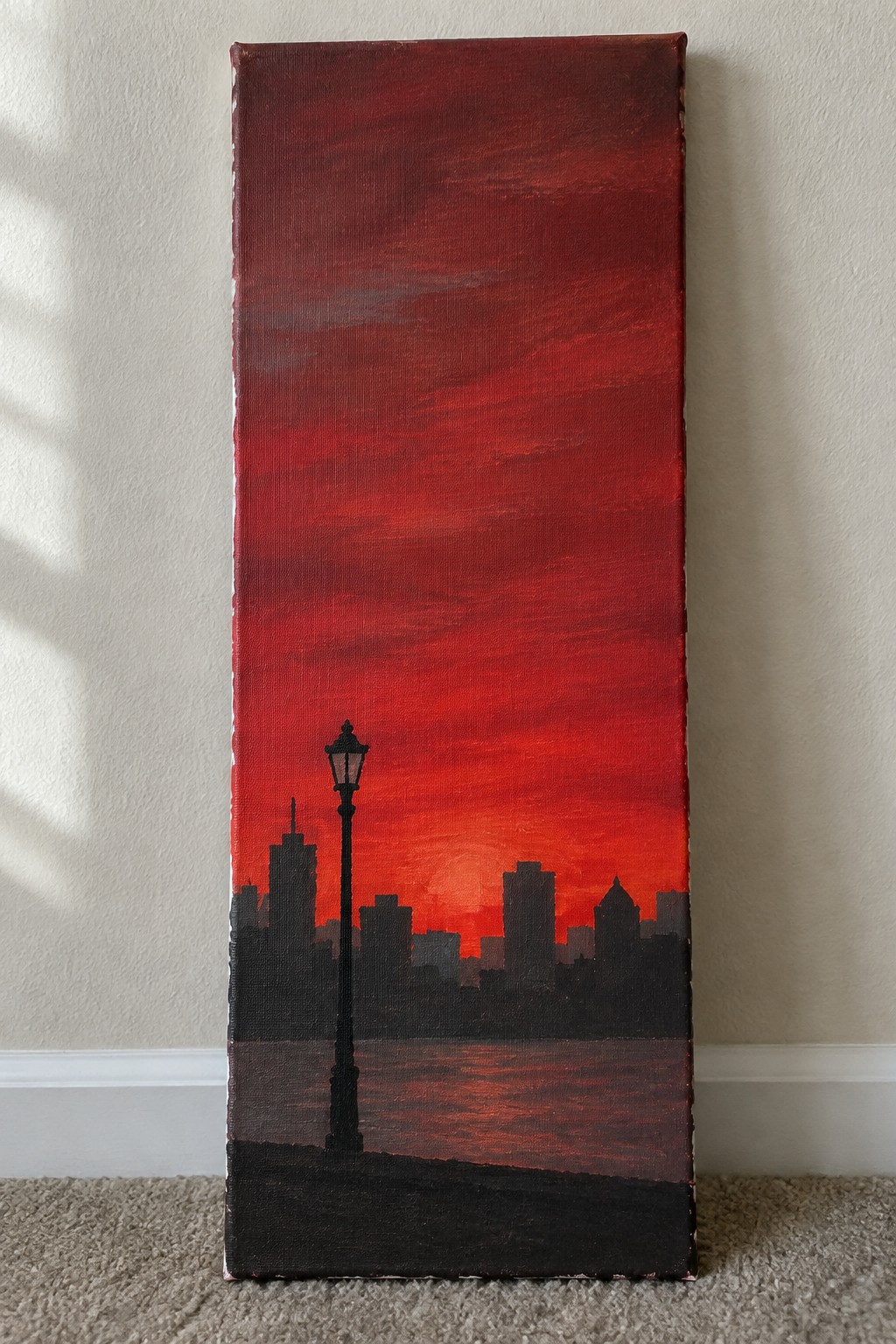 Tall canvas painting of black city skyline and lamppost under red sunset sky over water