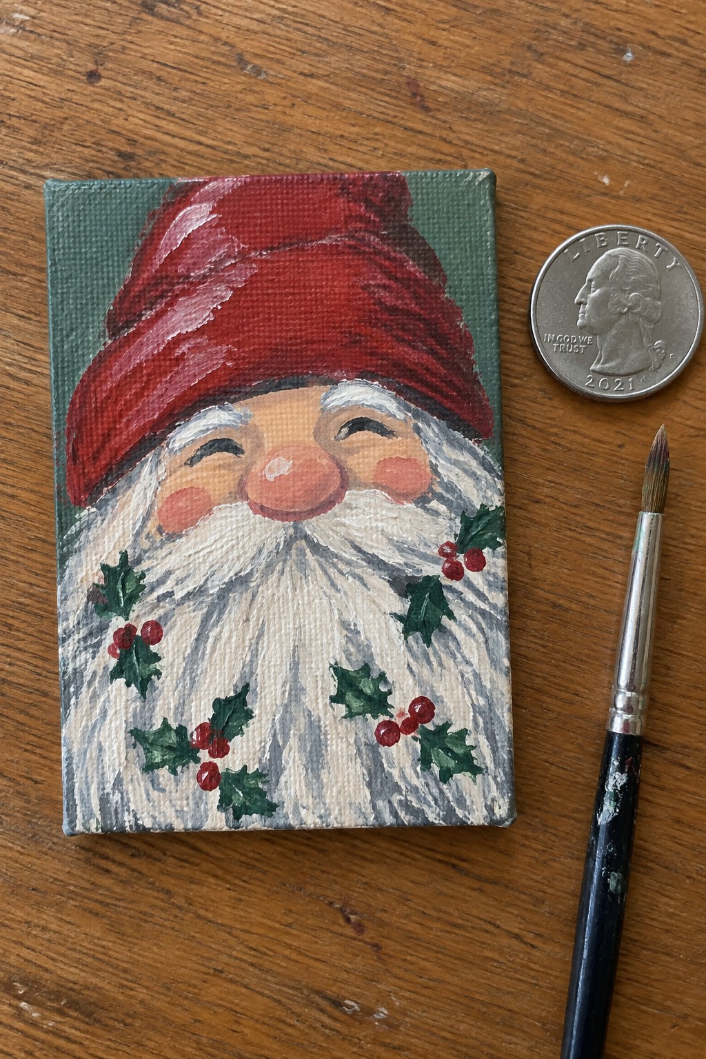 Small canvas painting of gnome in red hat with holly beside quarter and brush on wood