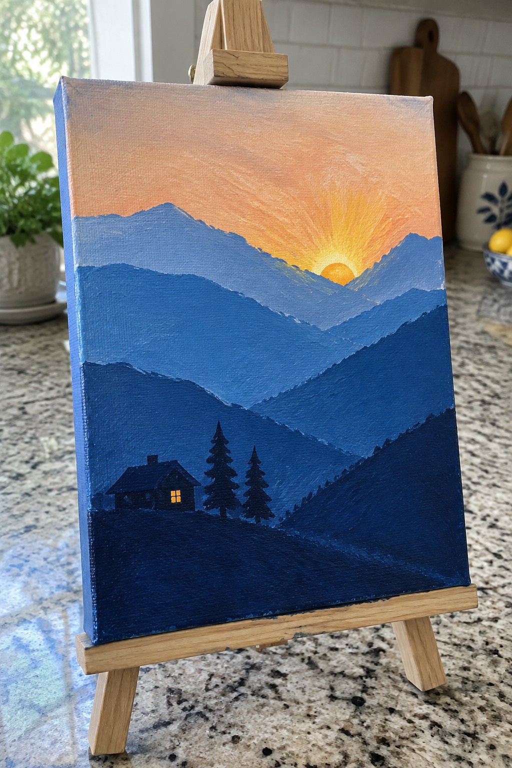 Painting on easel of sunset over layered blue mountains with cabin and pines.