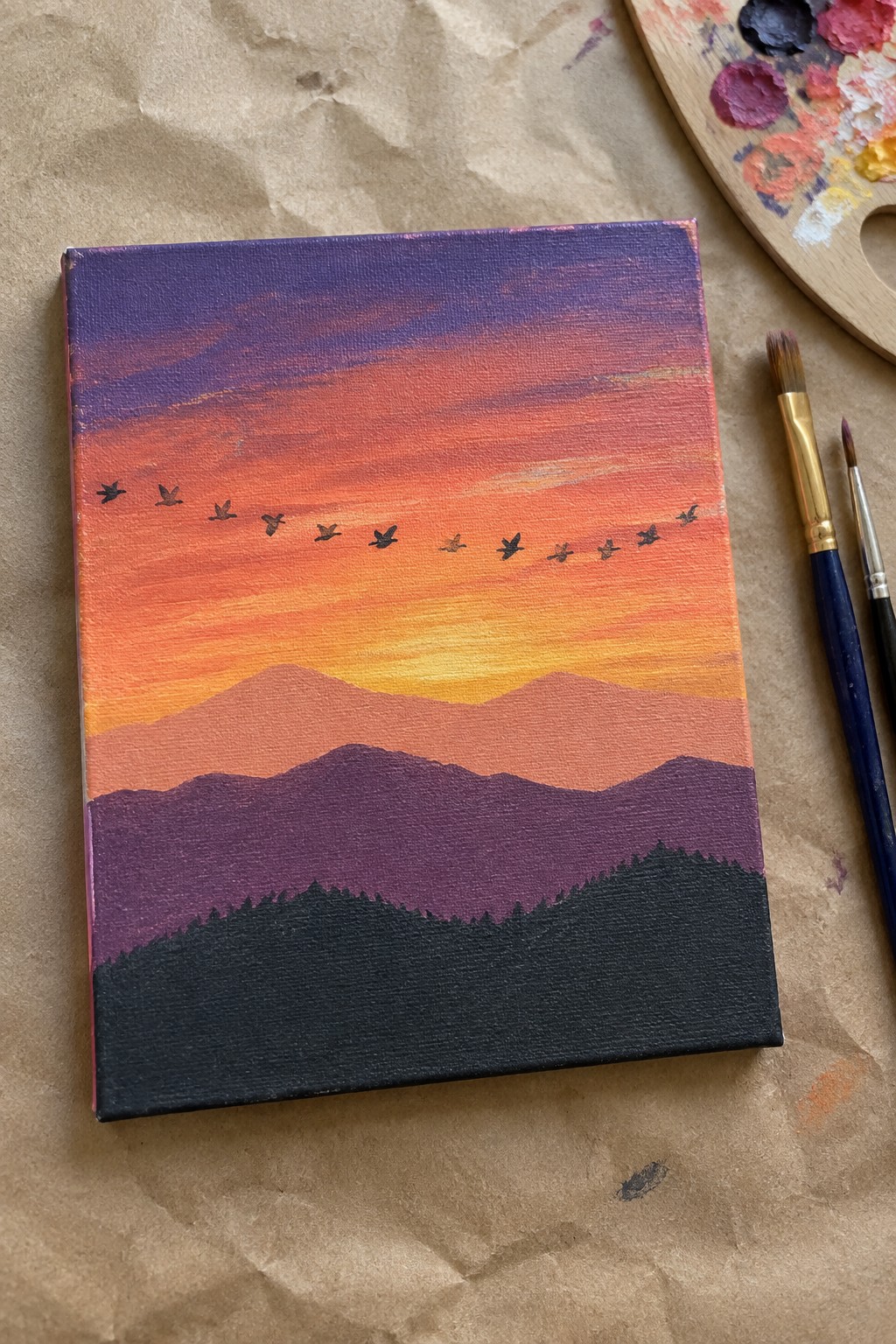 Canvas painting of gradient sunset over silhouetted mountains with flying birds and art supplies