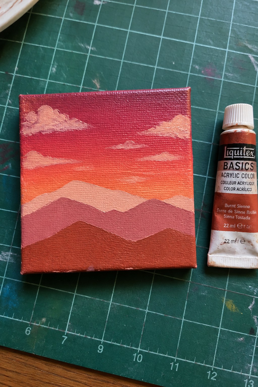 Small canvas with layered red-orange mountains beside burnt sienna acrylic paint tube on green mat.