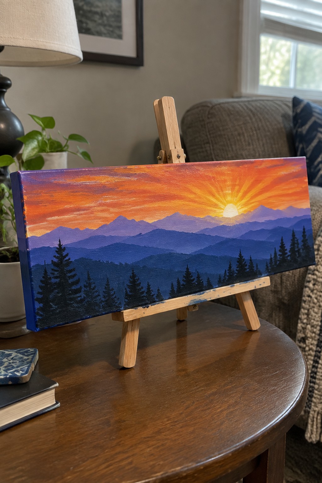 Vivid sunset painting of mountains and pine trees on small wooden easel.