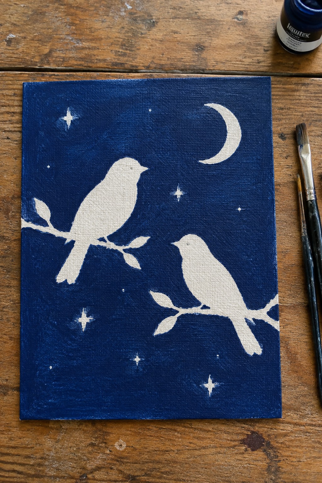 Dark blue canvas with two white birds, crescent moon and stars beside paintbrushes on wood.