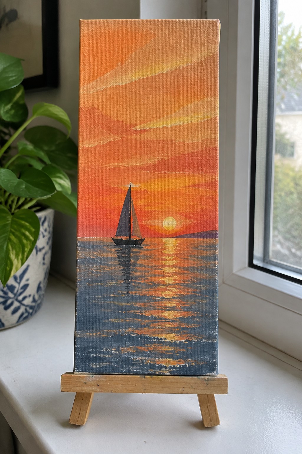 Vertical canvas on wooden easel showing sailboat against vibrant orange sunset over reflective sea.
