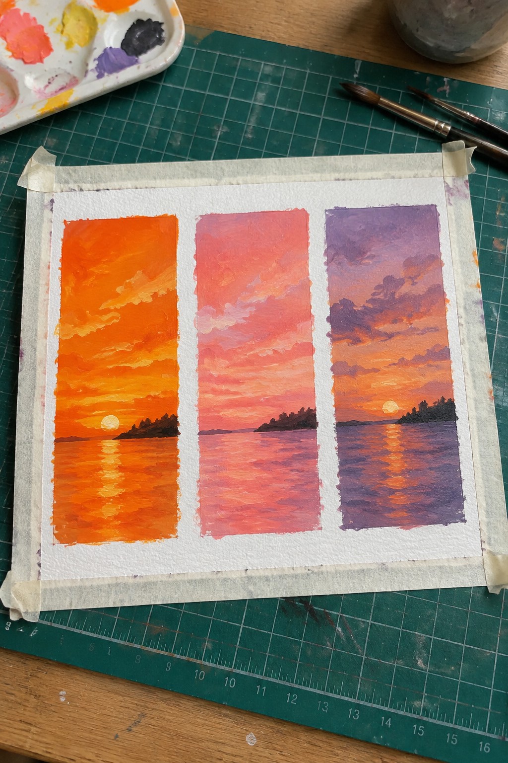 Three vibrant sunset paintings taped to green mat beside palette and brushes.