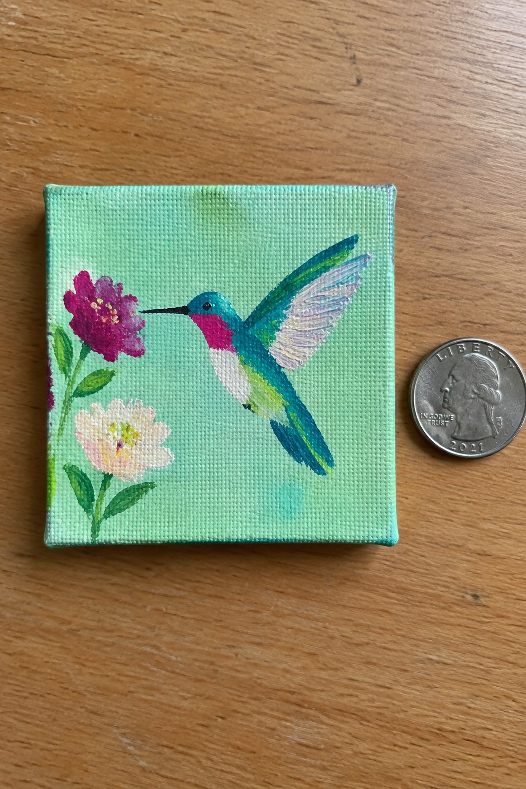 Miniature hummingbird painting on green canvas beside a 2021 US quarter on wood