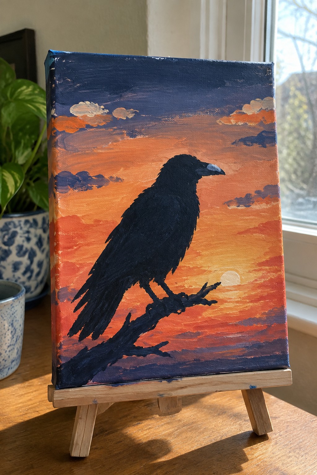 Painting of black raven on branch against vibrant orange-red sunset sky on wooden easel