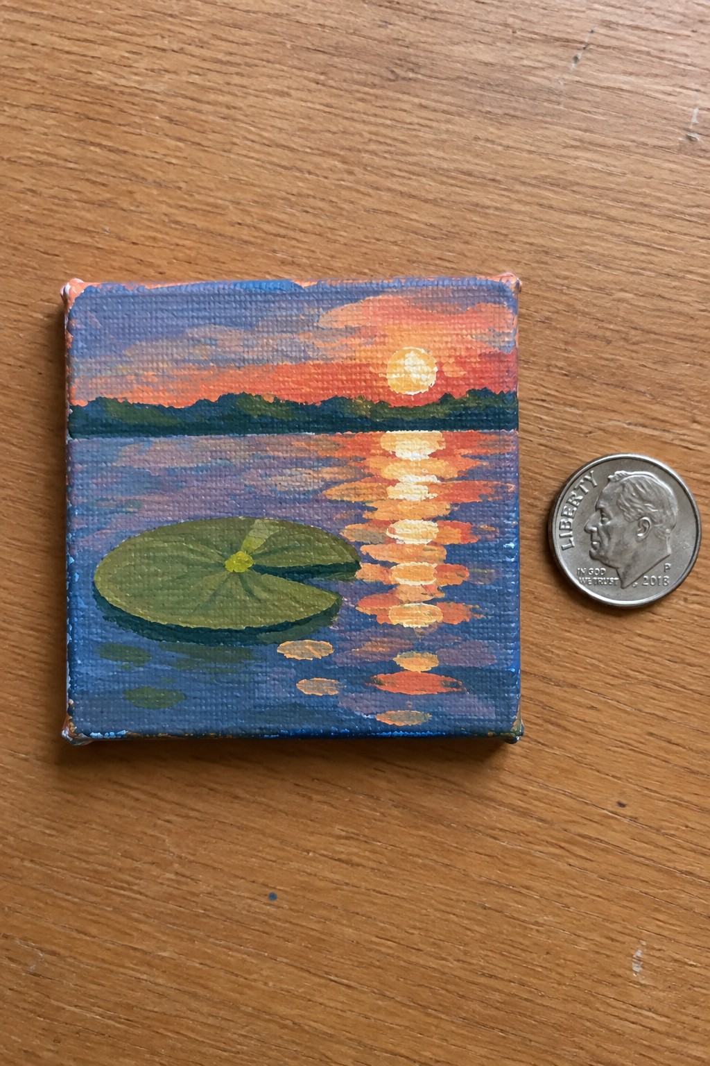 Miniature sunset painting with sun reflection and green lily pad beside 2018 dime.
