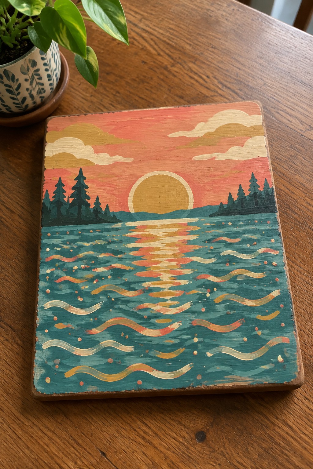 Sunset painting on wood with orange sky, yellow sun, pine trees, and wavy water.