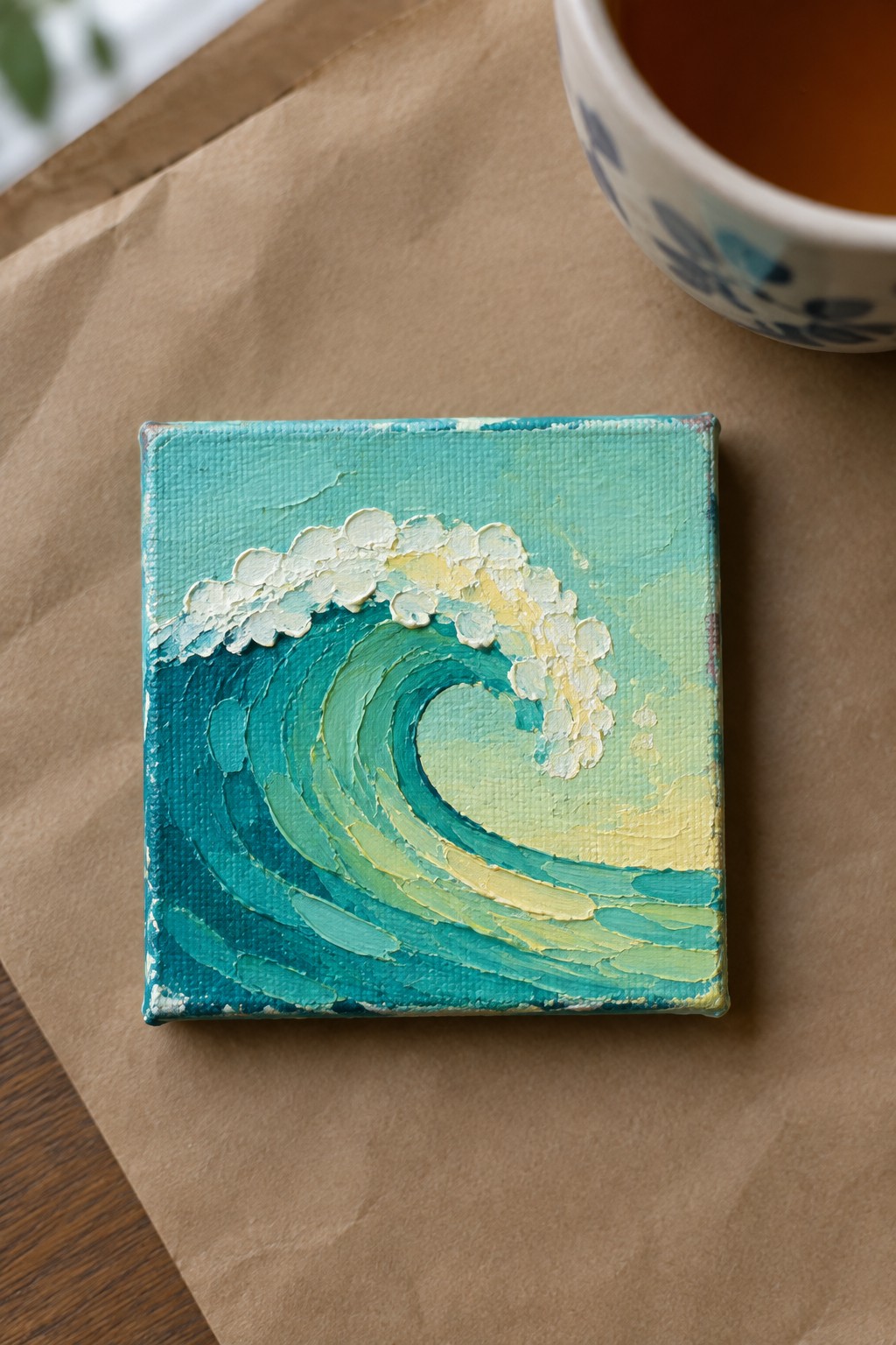 Small square canvas with curling teal wave painting beside blue-patterned cup on brown paper.