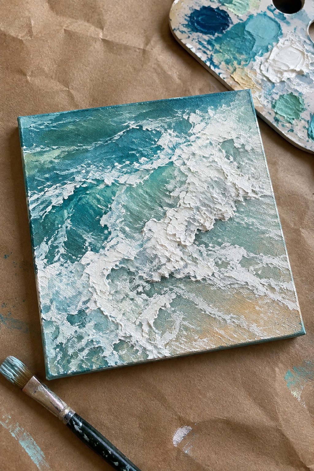 Textured turquoise wave painting on canvas beside paint palette and brush on brown paper