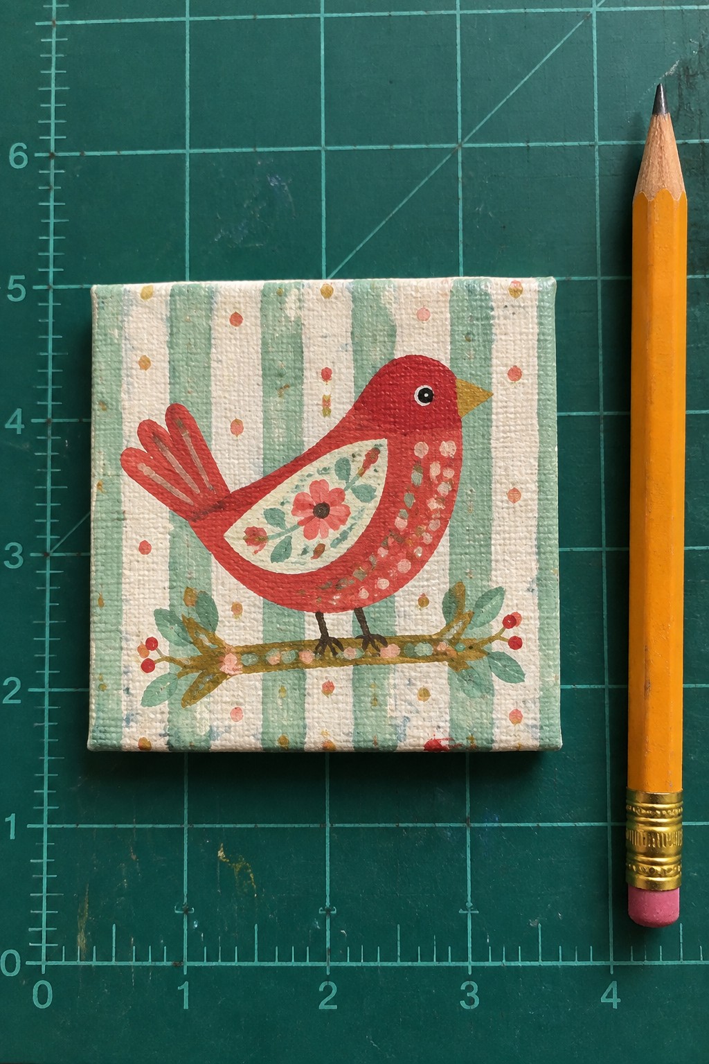 Miniature red bird painting on canvas beside yellow pencil on green grid mat