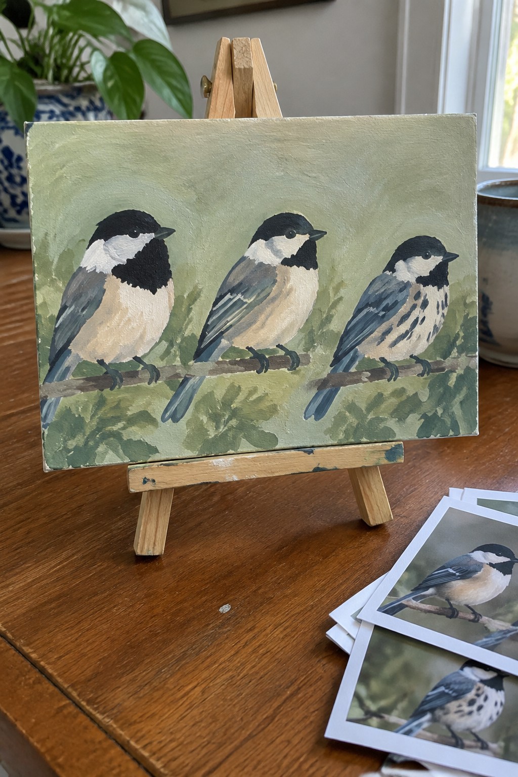 Acrylic painting of three chickadees on branches on a small canvas displayed on a wooden easel.