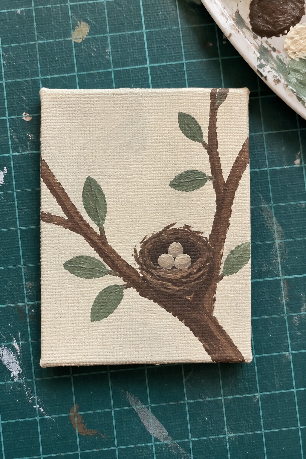 A small acrylic painting on canvas showing a brown tree branch with green leaves, a nest, and three eggs on an off-white background, placed on a green cutting mat.