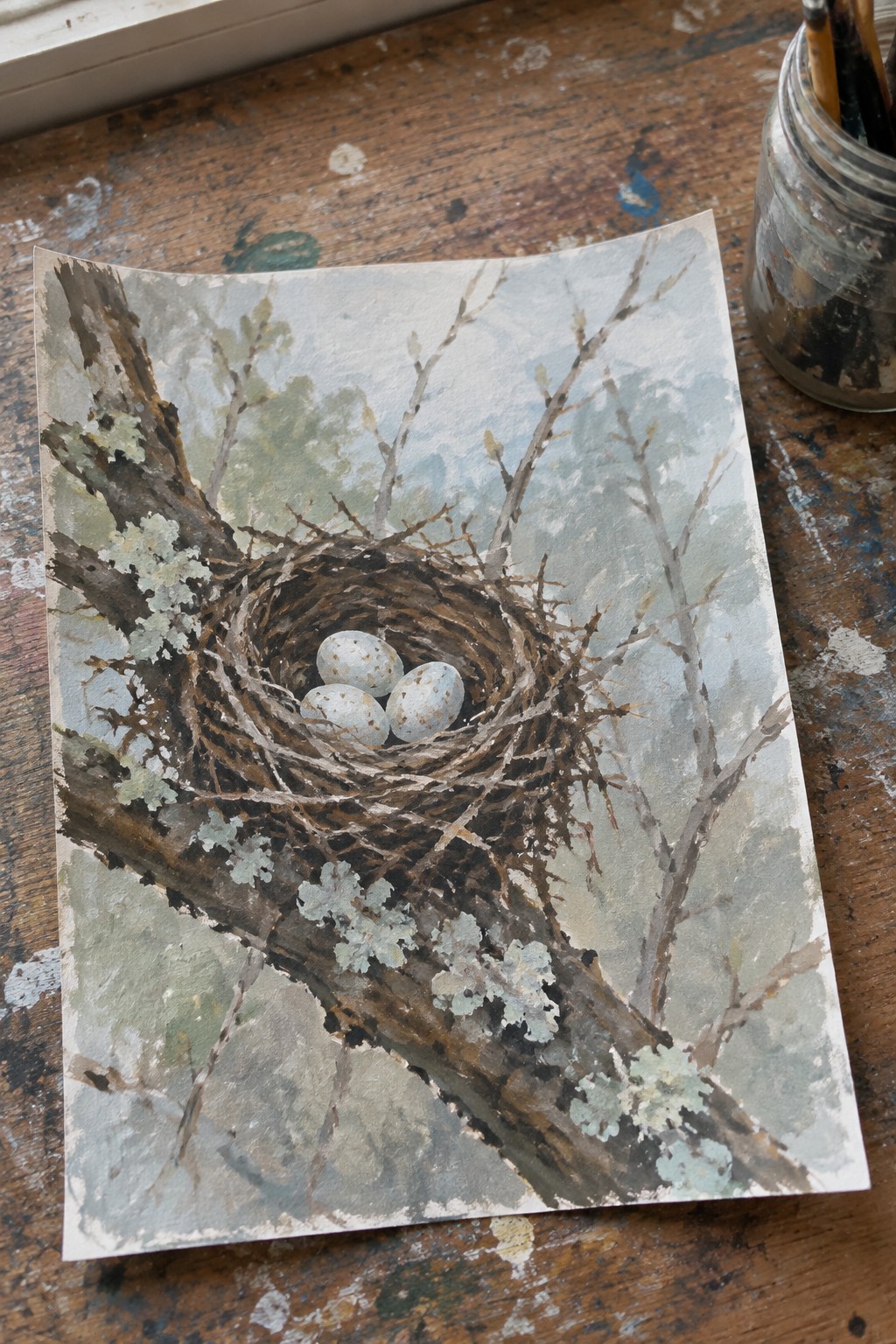 Watercolor painting of bird nest with three eggs on lichen-covered branch on wood table