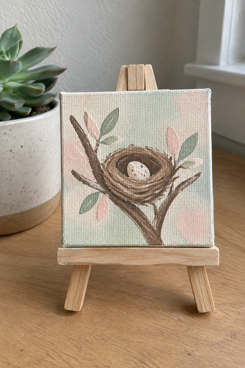 Small canvas on easel shows bird nest with egg amid pastel leaves.