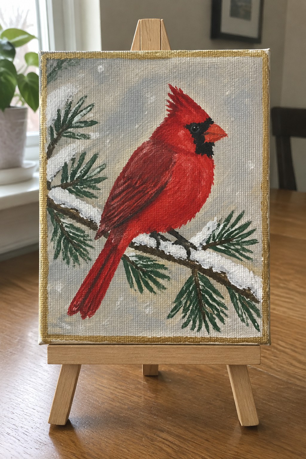 An acrylic painting of a red cardinal perched on a snowy pine branch inside a gold frame on an easel.