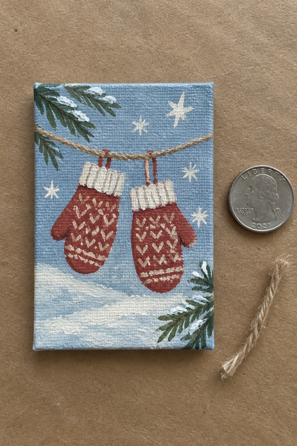 Two red mittens on twine in snowy painted scene on canvas next to quarter.