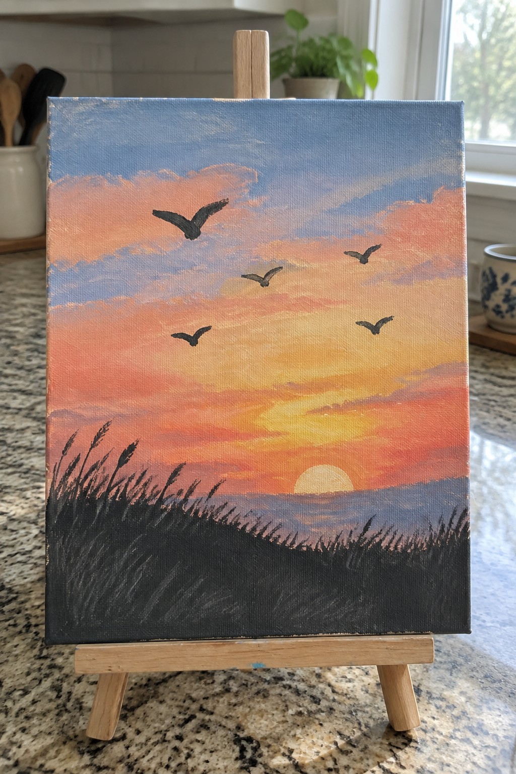 Sunset painting on easel with orange-pink sky, flying birds, and dark grass silhouette