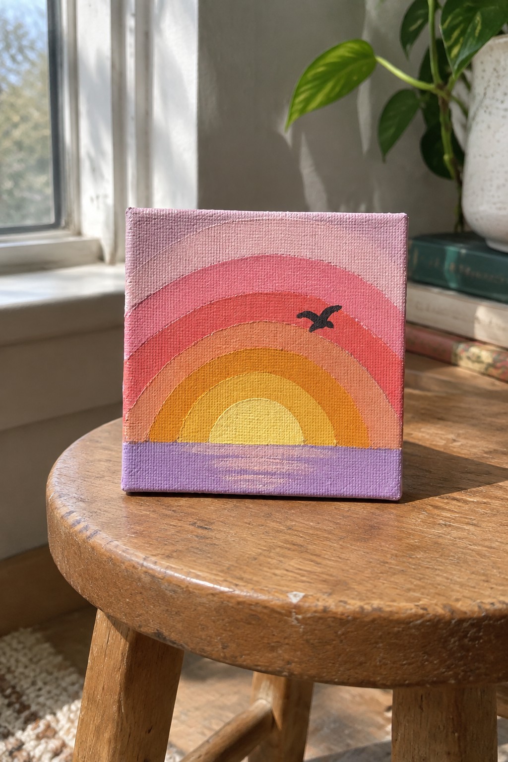 Small canvas rainbow painting with pink-orange arcs, yellow sun, bird on wooden stool