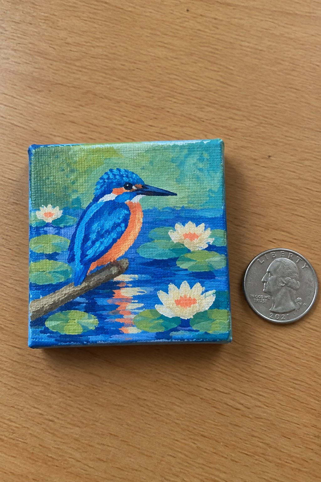 Small painting of blue kingfisher on branch above water lilies beside quarter coin.