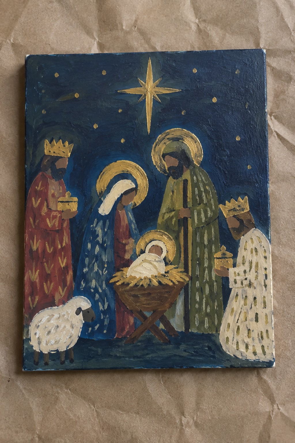 Folk art nativity painting with crowned kings, halos, manger, baby, sheep under golden star.