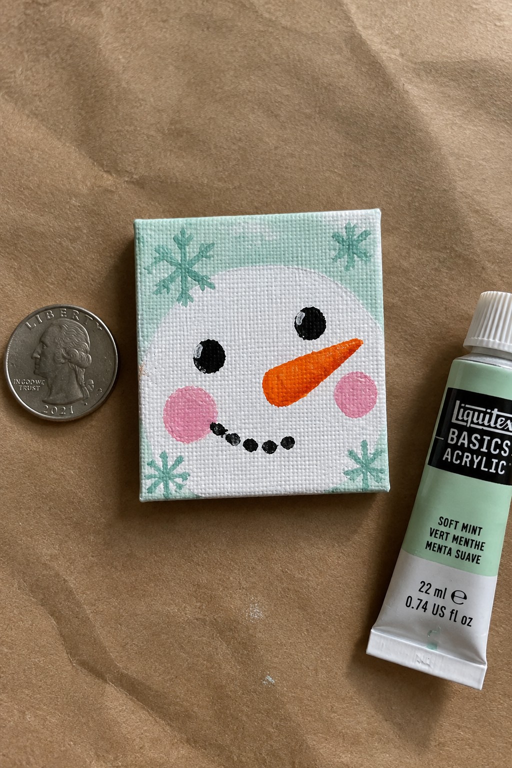 Snowman canvas painting beside US quarter and soft mint acrylic paint tube on brown paper