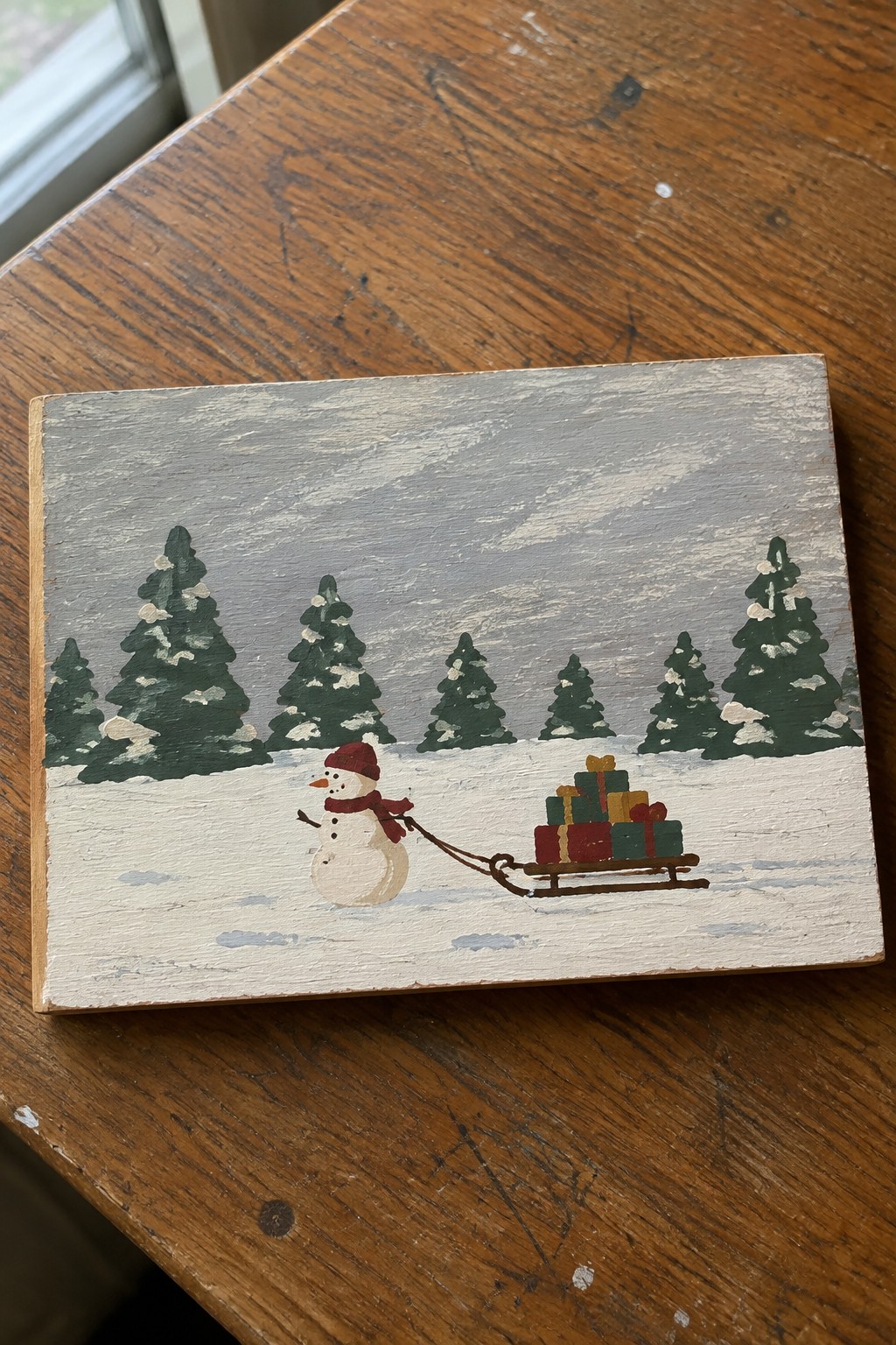 Folk painting of snowman in red hat pulling gift-filled sled past snowy pines