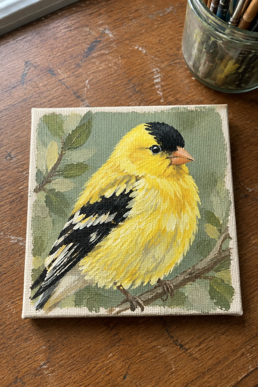 Yellow bird painting on canvas atop wooden table beside jar of brushes