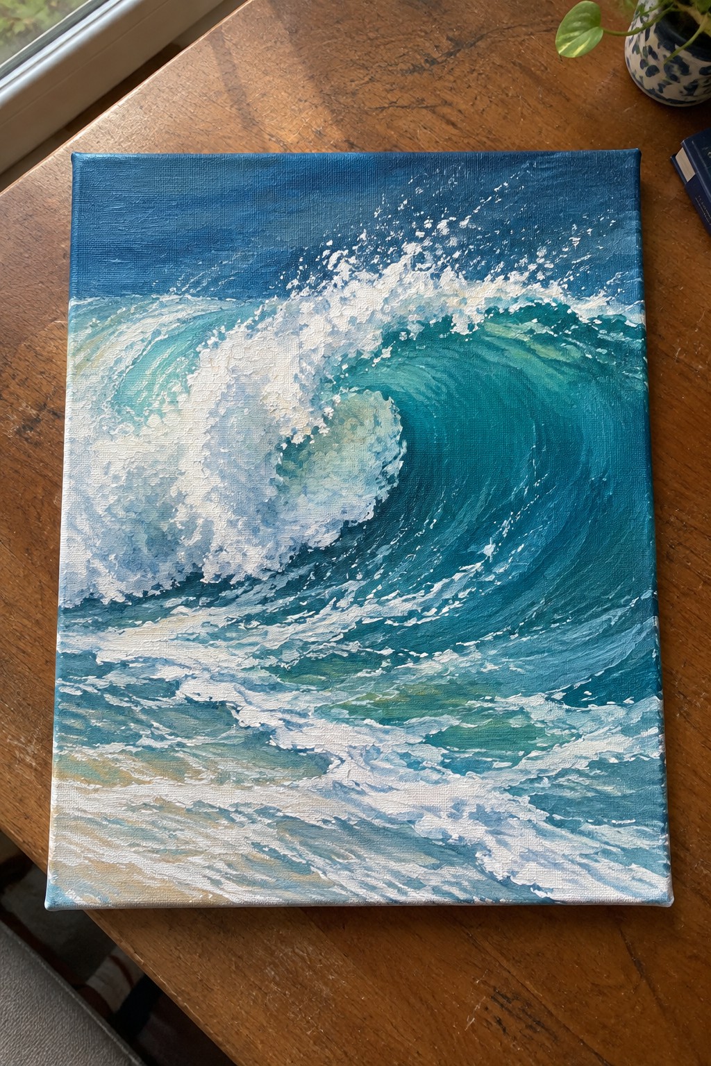 Painting of curling teal wave with white foam on wooden table beside plant