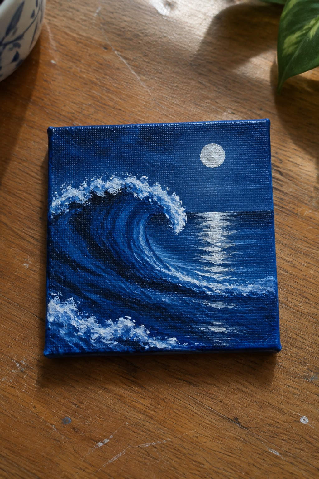 A small square acrylic painting of a dark blue curling ocean wave at night with a full moon and its reflection on the water.