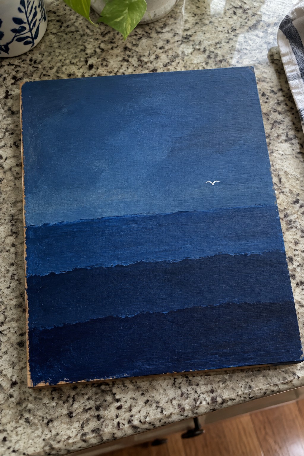 Abstract blue seascape painting with layered waves and small white bird on granite counter