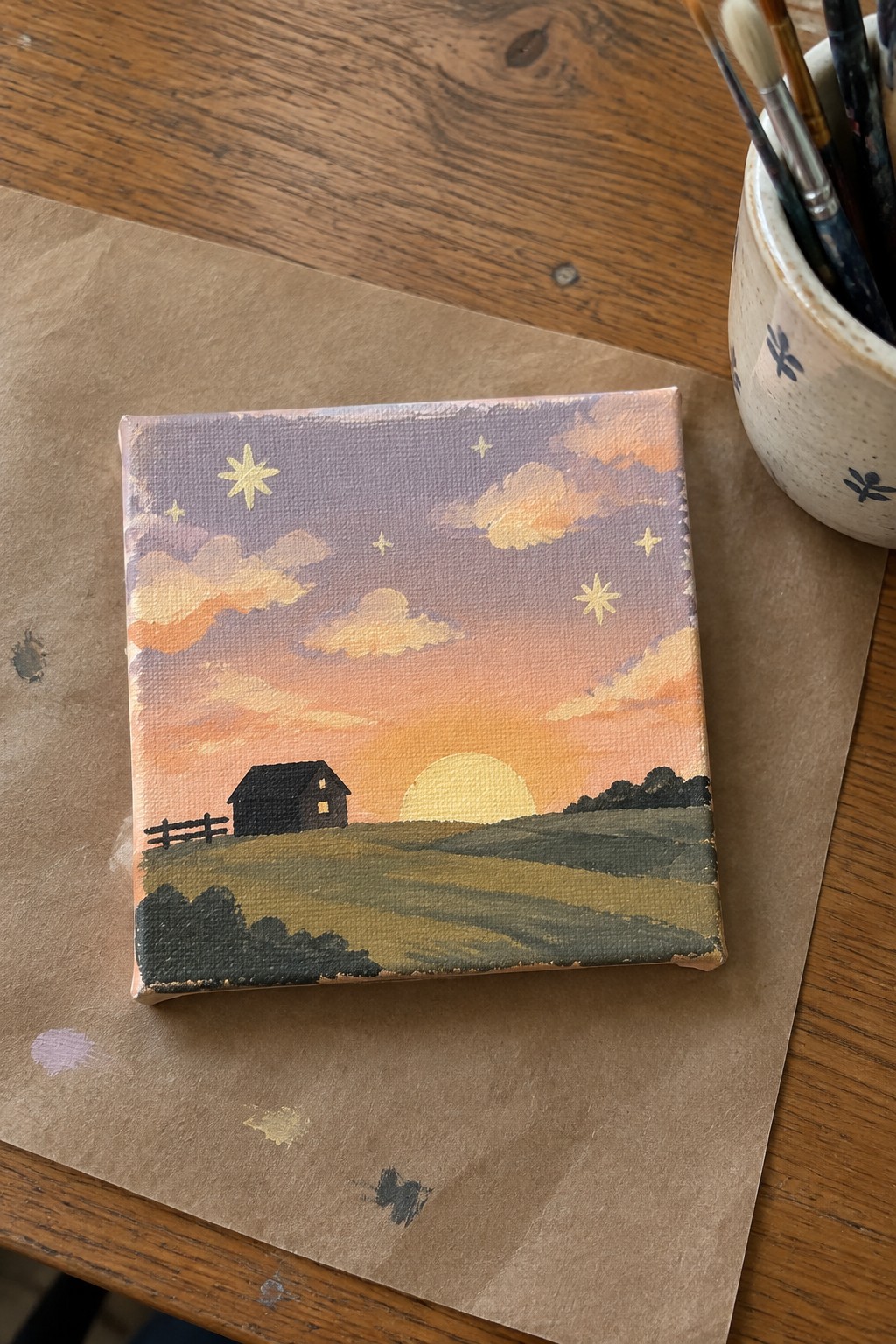 Mini canvas sunset painting of barn, fields, sun, clouds and stars on table